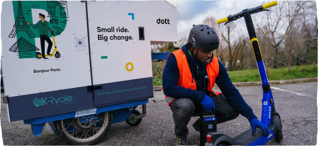 A man wearing a helmet and reflective clothing crouches down next to a Dott to repair it.